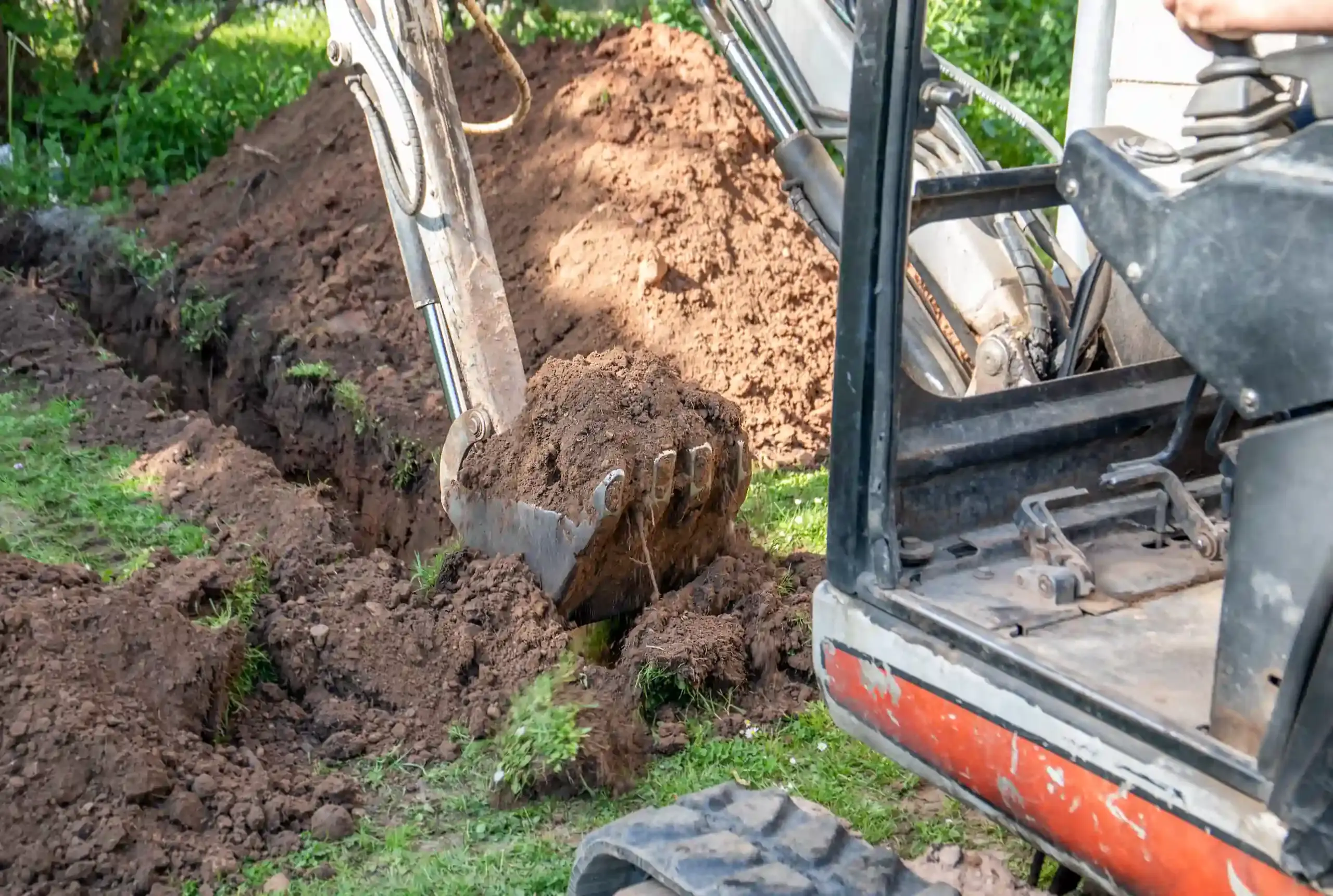 Minibagger hebt einen Graben in einer Rasenfläche aus, der Löffel führt Erdreich zum Aushubhaufen