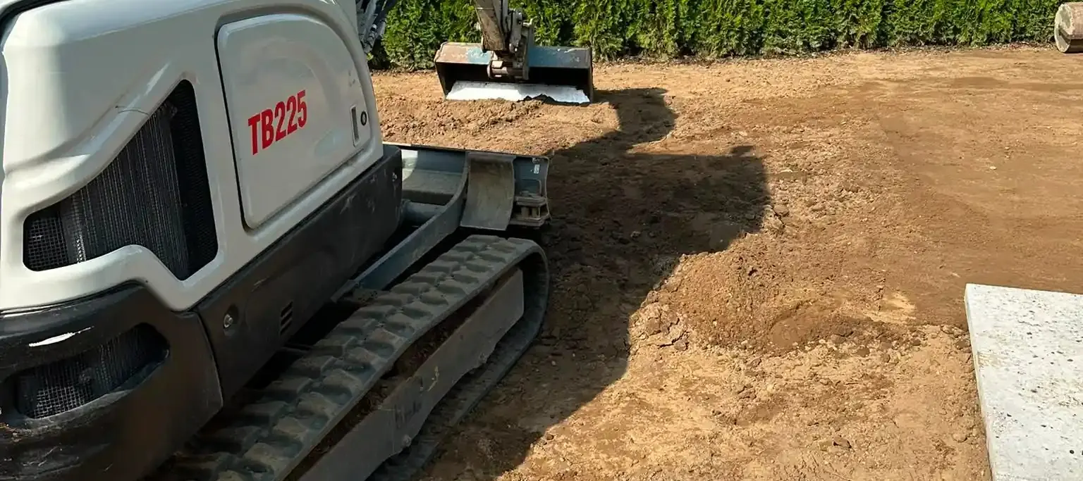 Minibagger zieht mit dem Löffel das Erdplanum ab, dahinter eine Thujahecke